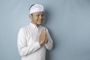 Smiling young Balinese man wearing udeng or traditional headband and white shirt gesturing greeting or namaste isolated over blue background photo