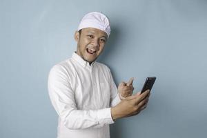 A portrait of a happy Balinese man is smiling and holding his smartphone wearing udeng or traditional headband and white shirt isolated by a blue background photo