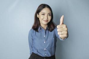 una mujer asiática emocionada con una camisa azul da un gesto de aprobación con la mano hacia arriba, aislada por un fondo azul foto