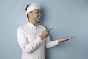 Excited Balinese man wearing udeng or traditional headband and white shirt pointing at the copy space beside him, isolated by blue background photo