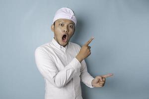 Shocked Balinese man wearing udeng or traditional headband and white shirt pointing at the copy space beside him, isolated by blue background photo