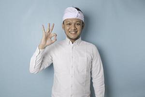 Excited Balinese man wearing udeng or traditional headband and white shirt giving an OK hand gesture isolated by a blue background photo