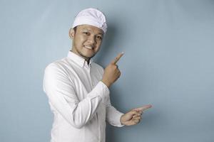 Excited Balinese man wearing udeng or traditional headband and white shirt pointing at the copy space beside him, isolated by blue background photo