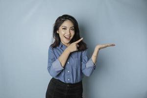 Excited Asian woman wearing blue shirt pointing at the copy space beside her, isolated by blue background photo