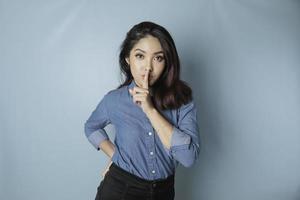 Portrait of a young Asian woman tell be quiet, shushing with serious face, hush with finger pressed to lips, isolated by blue background photo