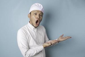 Shocked Balinese man wearing udeng or traditional headband and white shirt pointing at the copy space beside him, isolated by blue background photo