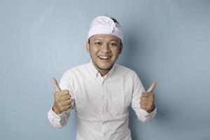 Excited Balinese man wearing udeng or traditional headband and white shirt gives thumbs up hand gesture of approval, isolated by blue background photo
