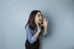 joven mujer hermosa con una camisa azul gritando y gritando fuerte a un lado con una mano en la boca. concepto de comunicación foto