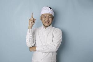 Excited Balinese man wearing udeng or traditional headband and white shirt pointing at the copy space upside him, isolated by blue background photo