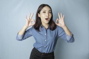 Excited Asian woman wearing a blue shirt giving an OK hand gesture isolated by a blue background photo