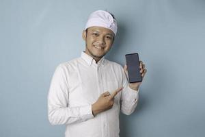Excited Balinese man wearing traditional headband or udeng and white shirt pointing at the copy space on his smartphone, isolated by blue background photo