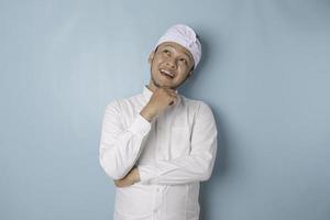 Portrait of a thoughtful young Balinese man wearing udeng or traditional headband and white shirt looking aside while his finger on his chin isolated over blue background photo