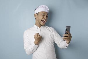 A young Balinese man with a happy successful expression wearing udeng or traditional headband and white shirt holding smartphone isolated by blue background photo