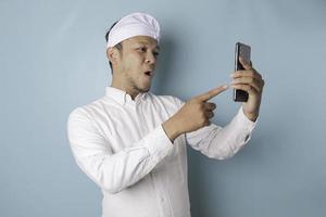 hombre balinés sorprendido con udeng o diadema tradicional y camisa blanca sosteniendo su teléfono inteligente, aislado de fondo azul foto