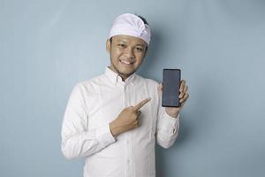 Excited Balinese man wearing traditional headband or udeng and white shirt pointing at the copy space on his smartphone, isolated by blue background photo