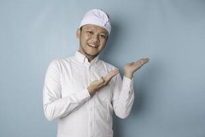 Excited Balinese man wearing udeng or traditional headband and white shirt pointing at the copy space beside him, isolated by blue background photo