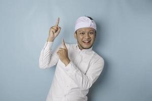 Excited Balinese man wearing udeng or traditional headband and white shirt pointing at the copy space upside him, isolated by blue background photo