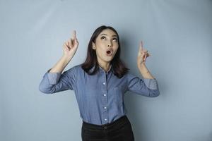 Shocked Asian woman wearing blue shirt pointing at the copy space upward, isolated by blue background photo