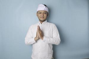Smiling young Balinese man wearing udeng or traditional headband and white shirt gesturing greeting or namaste isolated over blue background photo