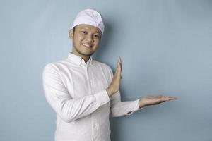 Excited Balinese man wearing udeng or traditional headband and white shirt pointing at the copy space beside him, isolated by blue background photo