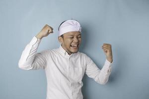 A young Balinese man with a happy successful expression wearing udeng or traditional headband and white shirt isolated by blue background photo