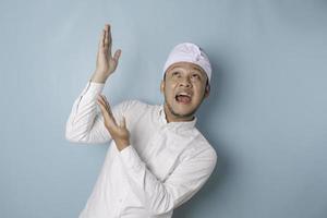 Excited Balinese man wearing udeng or traditional headband and white shirt pointing at the copy space upside him, isolated by blue background photo