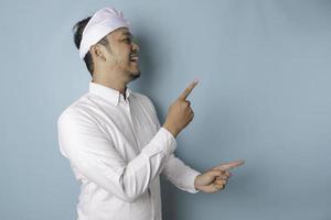 Excited Balinese man wearing udeng or traditional headband and white shirt pointing at the copy space beside him, isolated by blue background photo