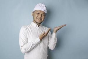 Excited Balinese man wearing udeng or traditional headband and white shirt pointing at the copy space beside him, isolated by blue background photo