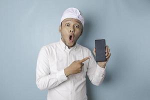 hombre balinés sorprendido con udeng o diadema tradicional y camisa blanca sosteniendo su teléfono inteligente, aislado de fondo azul foto