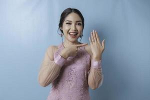 una joven asiática sonriente que lleva una kebaya moderna mostrando su anillo de compromiso aislado de fondo azul foto