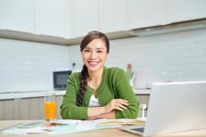 Smiling young woman sit at desk in kitchen working on laptop gadget, happy millennial girl manage finances pay household expenditures or taxes online, photo