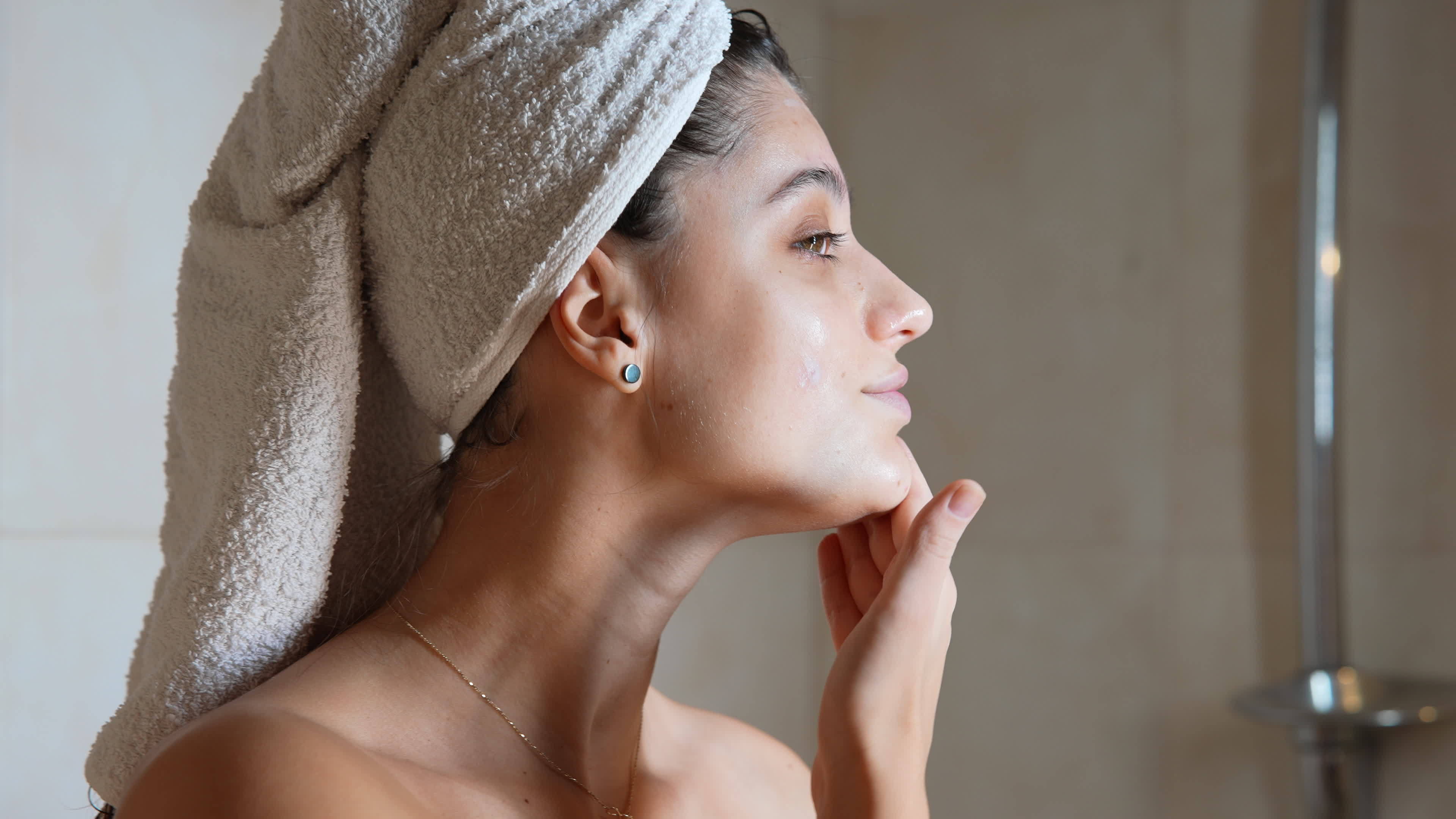 Woman with hair in towel smooths moisturizer on face after shower