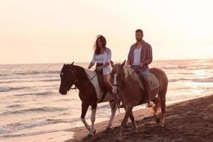 una pareja amorosa vestida de verano montando a caballo en una playa de arena al atardecer. mar y puesta de sol de fondo. enfoque selectivo foto