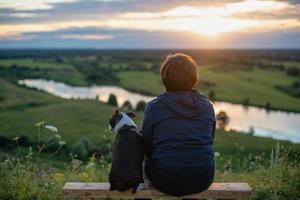 A boy and a dog sitting back from behind and looking at the setting sun in nature in autumn. boy embraces his pet on nature, concept of emotional health in harmony photo