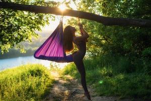 A young female gymnast is engaged in aerial yoga, using a combination of traditional yoga poses, pilates and dance using a hammock at sunset in nature. Healthy lifestyle. photo