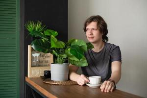 un adolescente positivo se sienta a la mesa en casa en la cocina con una taza de café en un vaso, se relaja y descansa por la mañana. foto