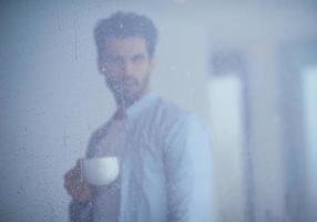 un joven relajado bebe el primer café de la mañana con gotas de lluvia en la ventana foto