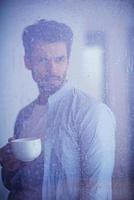 relaxed young man drink first morning coffee withh rain drops on window photo