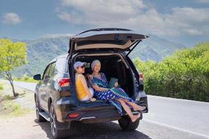 Two happy woman sitting at the car trunk on a road trip. photo