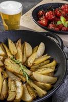 Roasted potatoes with rosemary in iron casserole and plate of confit tomatoes on dark background