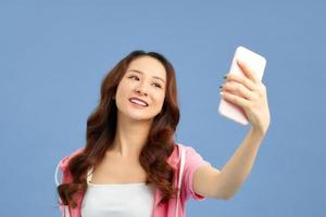 Portrait of cool cheerful girl having video-call with lover holding smart phone in hand, shooting selfie on front camera isolated on blue background. photo