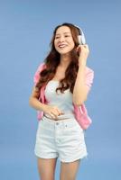 Energy Asian girl with headphones listening to music with closed eyes on blue background in studio. She wears white T-shirt, shorts. photo