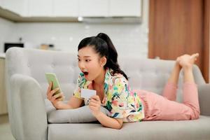 Image of optimistic excited young woman indoors at home using mobile phone holding credit card on sofa. photo
