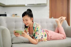 una joven asiática sonriente que usa una aplicación de teléfono móvil para jugar, relajarse en el sofá. foto