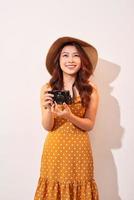 Portrait of cheerful smiling young woman taking photo with inspiration and wearing summer dress. Girl holding retro camera. Model posing on beige background in hat