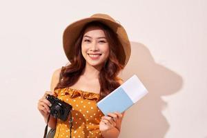 retrato de una joven feliz con sombrero sosteniendo una cámara y mostrando el pasaporte mientras se encuentra aislada sobre un fondo beige foto