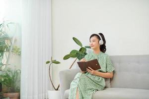 mage of focused asian woman using headphones and writing down notes while sitting on sofa at home