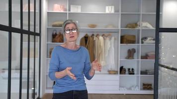 Woman with gray hair and glasses stands in front of a clothing store display talking to the viewer and gesturing with hands then crosses arms and smiles video