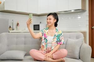 Pretty Young Asian Woman sits on sofa hand holding smartphone taking selfie photo