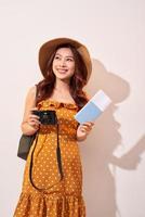 Portrait of a happy young woman in hat holding camera and showing passport while standing isolated over beige background photo